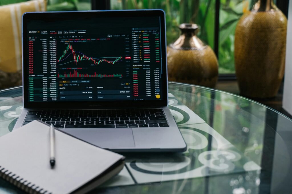 Laptop exibindo gráficos de ações e indicadores financeiros em tempo real, ao lado de um caderno e uma caneta sobre uma mesa de vidro.