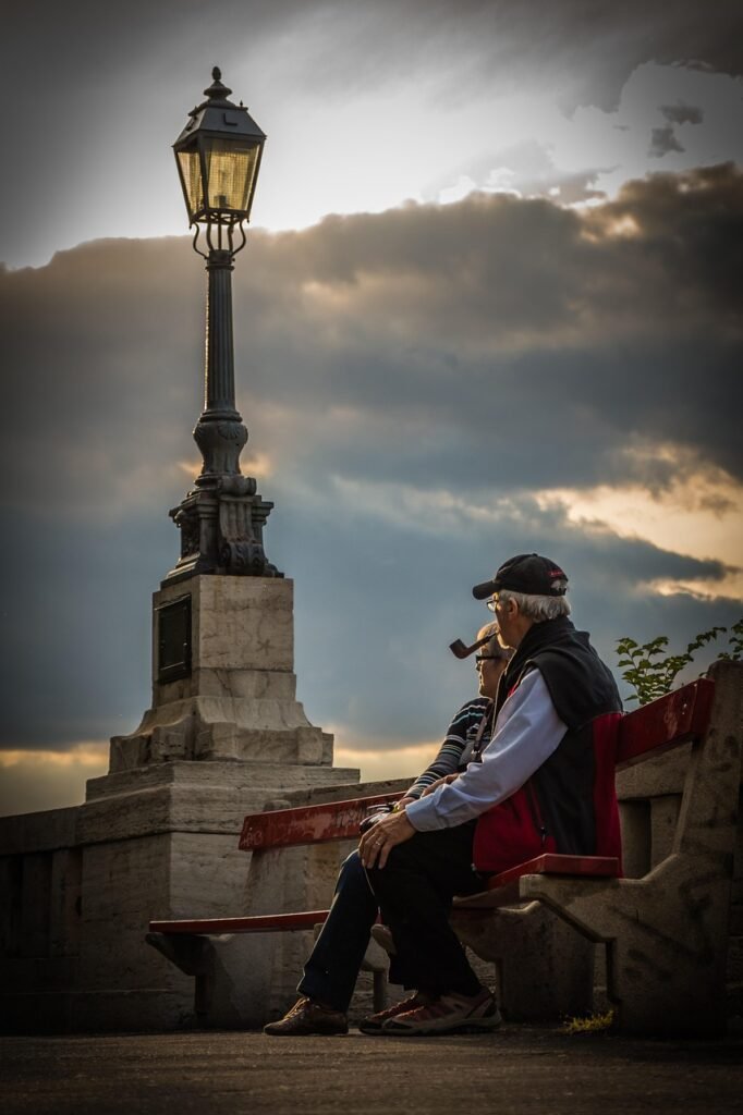 Casal idoso sentado em um banco ao lado de um grande poste de luz ornamental, sob um céu nublado ao entardecer; o homem à direita usa boné e segura um cachimbo.