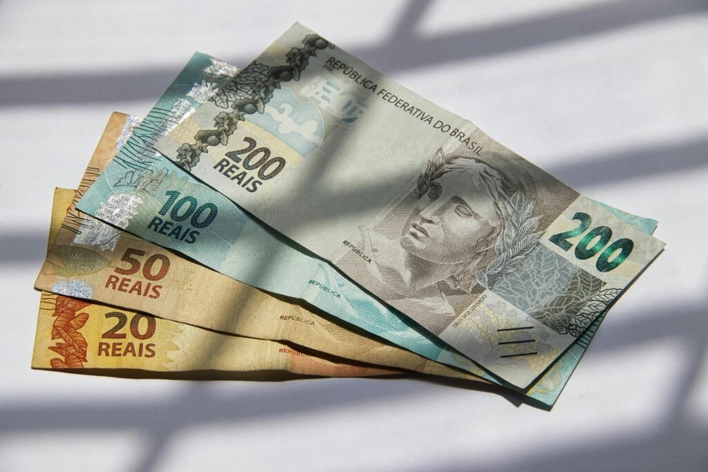 Notas de 20, 50, 100 e 200 reais do Brasil sobre uma superfície branca com sombras ao fundo.