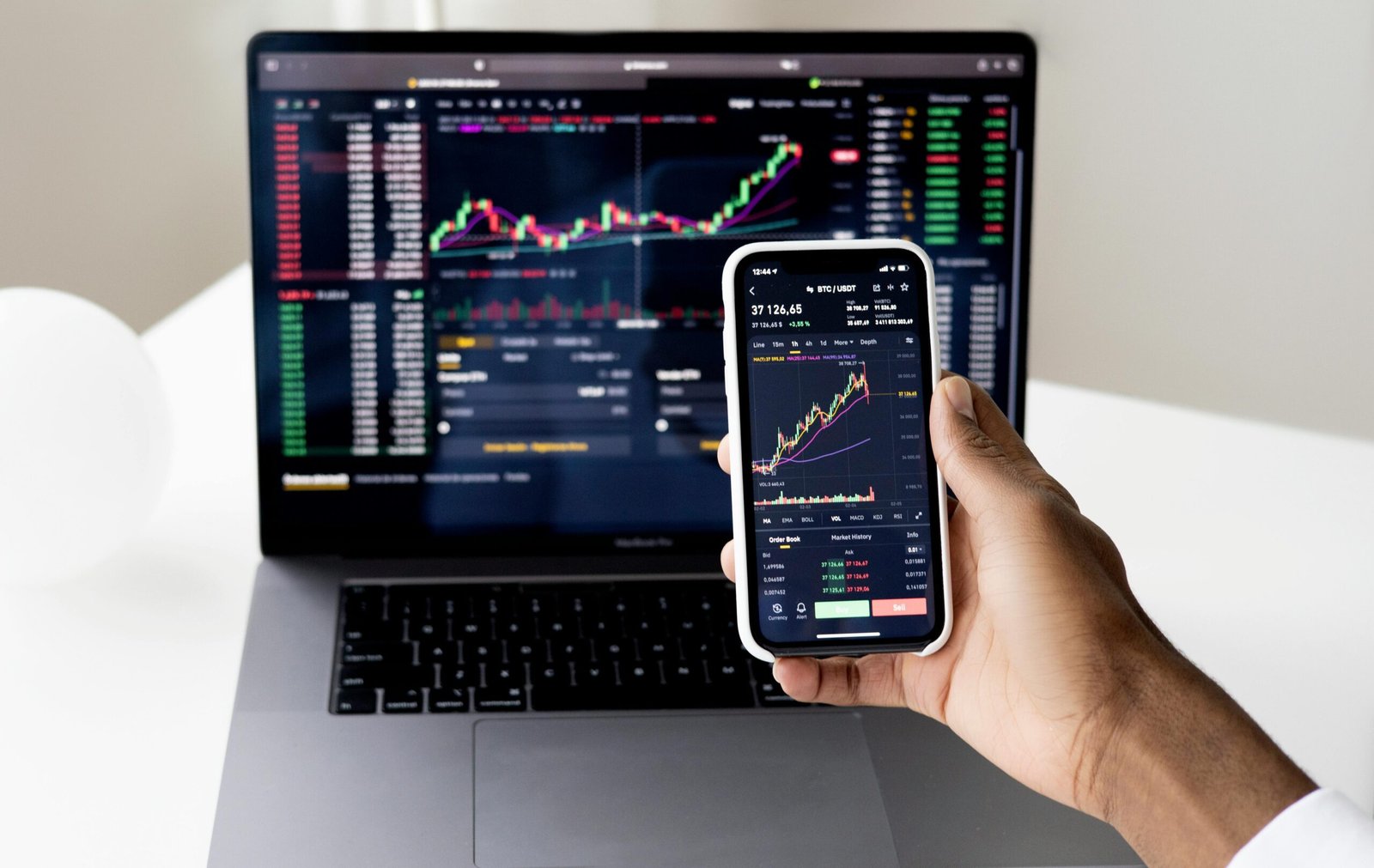 Mão segurando smartphone com gráficos de investimentos em fundos multimercado, com laptop ao fundo mostrando plataforma de trading com gráficos de candlestick e indicadores financeiros